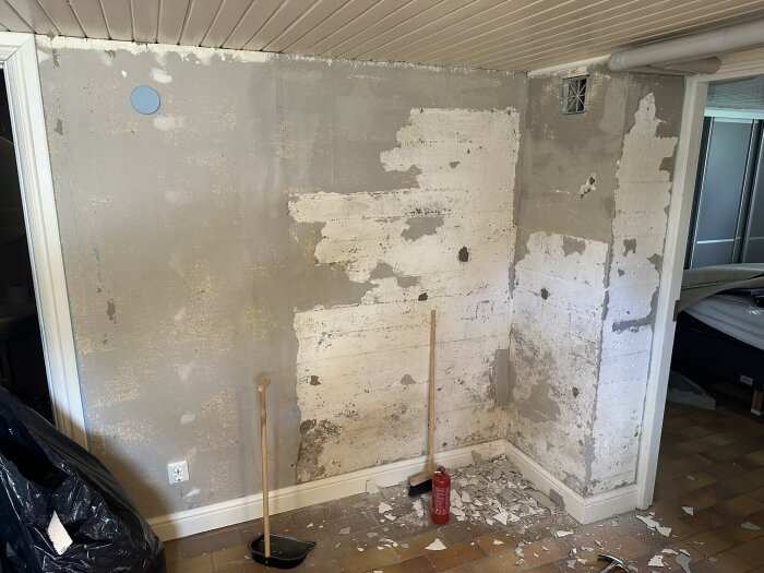 Vägg i källare med avflagad puts, synliga fuktfläckar och några verktyg på golvet. Ventilationsrör i hörnet och en brandsläckare är närvarande.