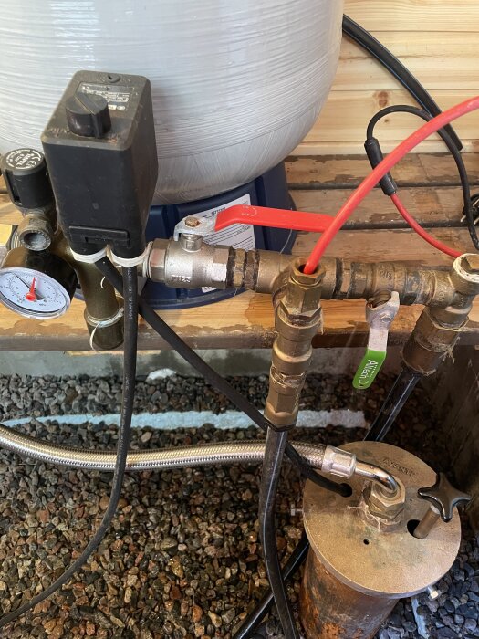 En kulventil med två handtag, ansluten till rör och en trycktank, omgiven av slangar och mätare, på en trävägg och grusbotten.