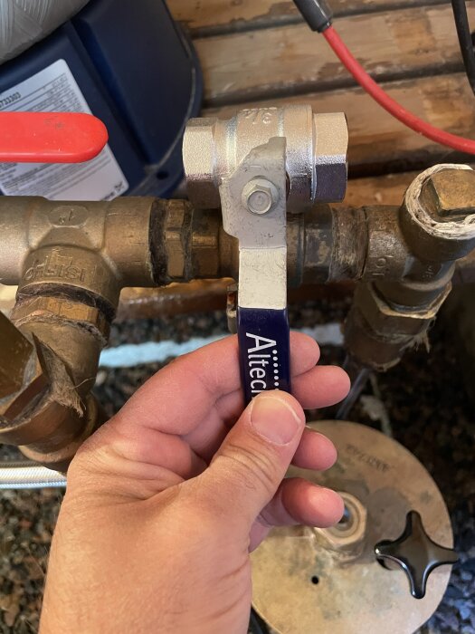 En hand håller i en metallisk kulventil med en blå spakhandtag märkt "Altech", monterad på ett kopparrör, med en röd ventil i bakgrunden.