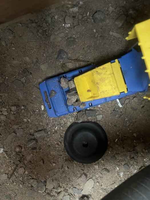 Blå och gul plastbit samt svart föremål ligger på grusig mark.