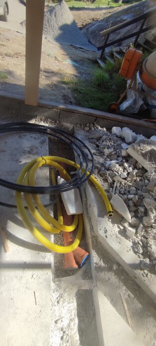 Betongplatta med utskuret spår för vatten- och avloppsrör, omgivande grus och isoleringsmaterial synligt, samt slangar och rör på plats.