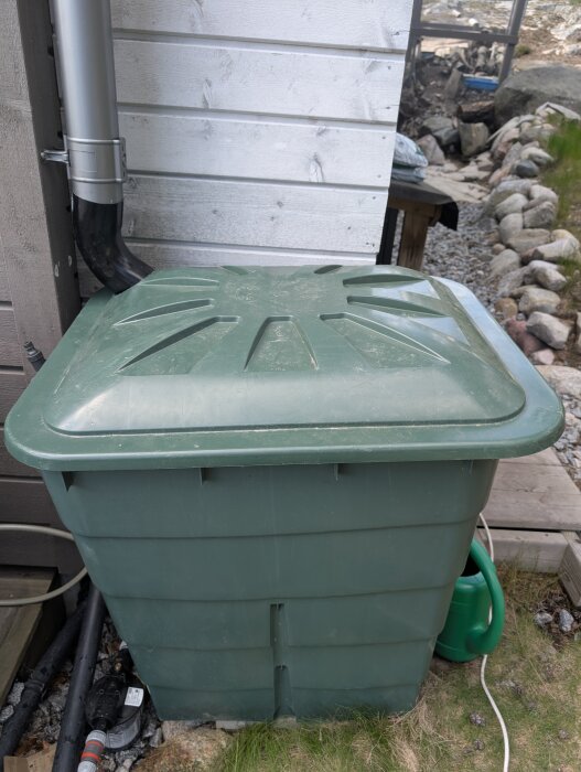 Grön vattentunna samlar regnvatten med kopplat stuprör vid vit husvägg, del av automatiskt bevattningssystem.