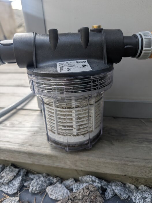 Automatiskt bevattningssystem med ett vattenfilter placerat på en träyta, anslutet med slangar och omgivet av stenar, för odlingslåda.