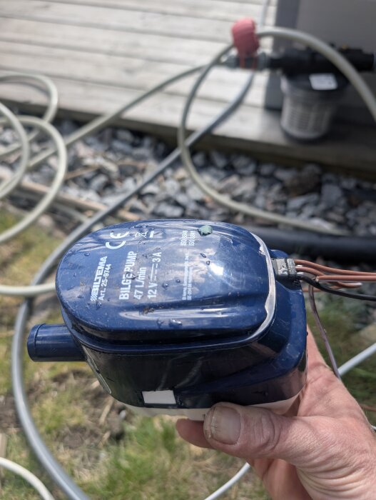 Hand håller en blå vattenpump till ett automatiskt bevattningssystem, med slangar i bakgrunden på gräsmatta vid altan.