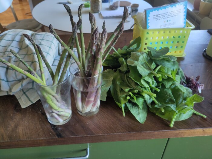 Färsk skördad sparris i glas och spenatblad på ett köksbord, omgiven av en kökshandduk och en gul plastkorg i bakgrunden.