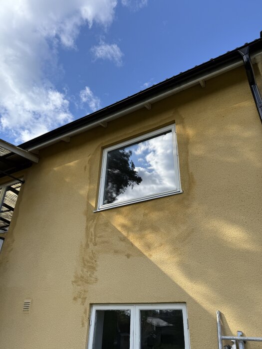 Putsad fasad med fönster och omgivande fläckar av annan färgnyans synliga mot en klarblå himmel.