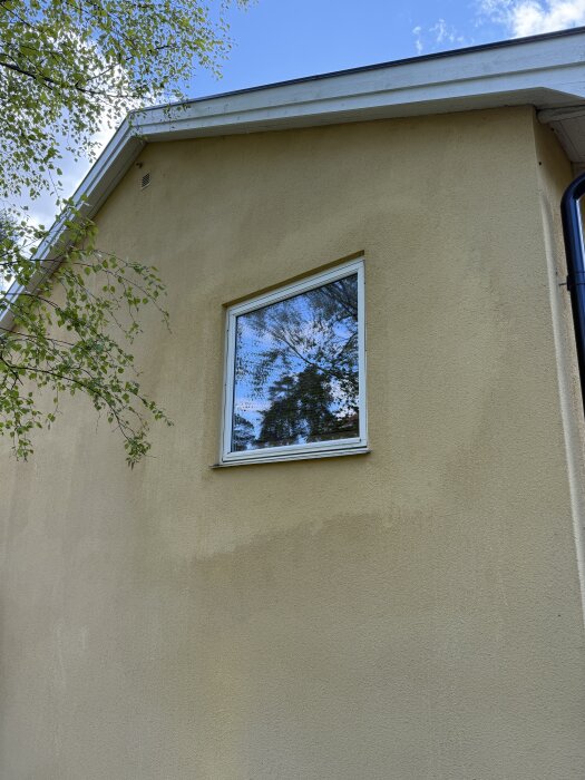 Putsad husfasad med ett fönster, äldre silikatfärg och områden med nyare akrylatfärg synliga runt fönstret, blå himmel i bakgrunden.