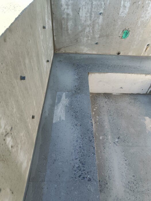 Betongyta som har slipats, med synliga märken och ojämnheter på golvet och bänken. Vägg med fästen och grönt märke syns i bakgrunden.