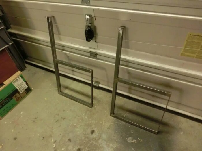 Svetsade metallkonsoler stående mot garagedörr, för hyllprojekt.