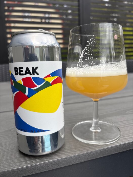 En ölburk med färgglad etikett bredvid ett glas med ljus, skummig öl står på ett bord utomhus.