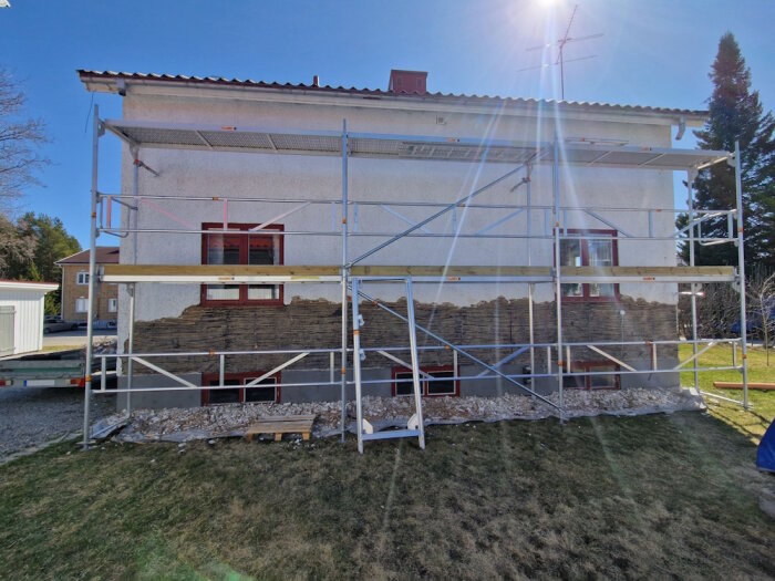 Ställning vid husfasad där puts avlägsnas, med gräsmatta i förgrunden och blå himmel. Solstrålar lyser mot väggen.