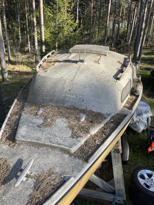 Smutsig segelbåt på land under tallar, täckt av barr och skräp, redo för rengöring med högtryckstvätt.
