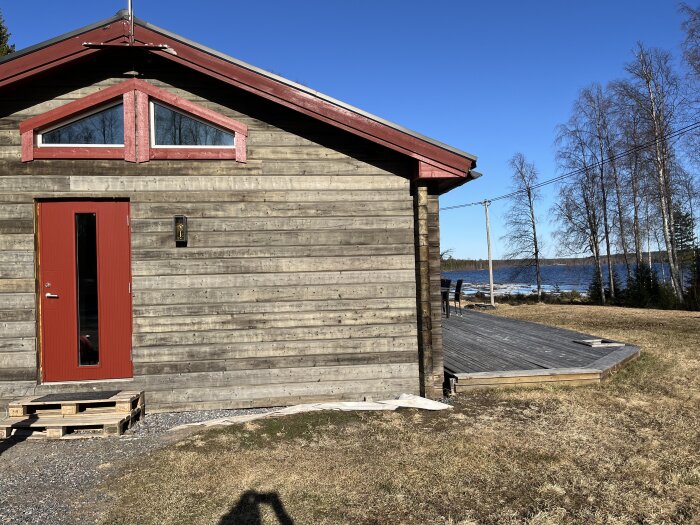 Trähus med röd ytterdörr, stor altan i tryckimpregnerat virke, soligt väder, utsikt över sjö och skog i bakgrunden.