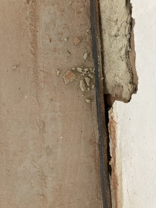 Golv med skumplastremsa bredvid murad vägg; glipor synliga mellan plastremsan och väggen, med puts som delvis borta och smulor på marken.