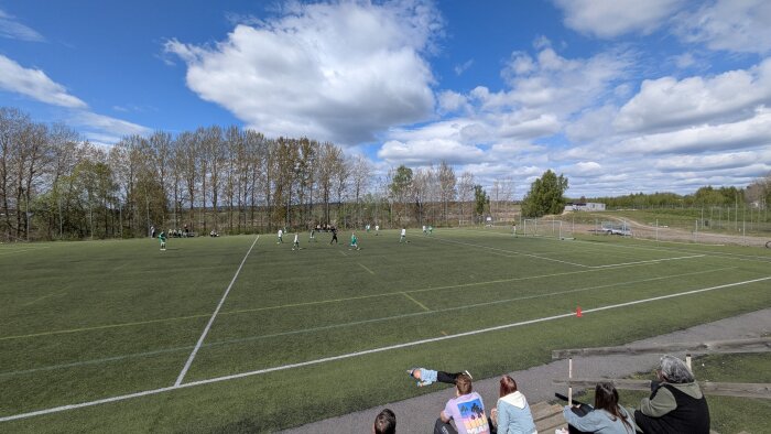 En fotbollsplan med spelare i gröna och vita tröjor, blå himmel och åskådare på läktaren.