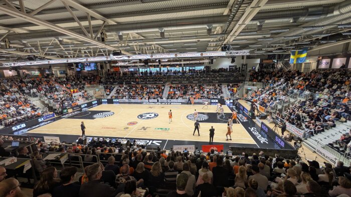 Basketmatch i en fullsatt arena med åskådare på läktarna och spelare i orange kläder på planen under sportsöndag.