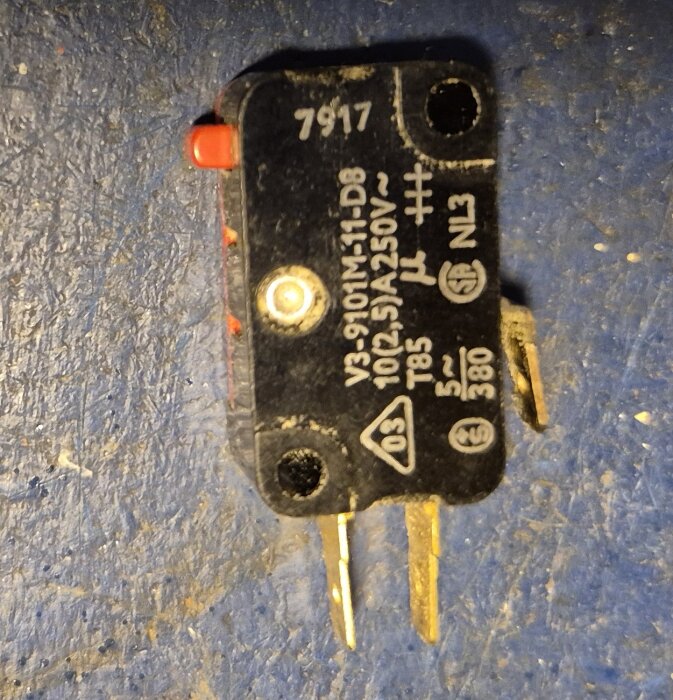 Svart mikrobrytare med märkningar och anslutningsstift mot blå yta.