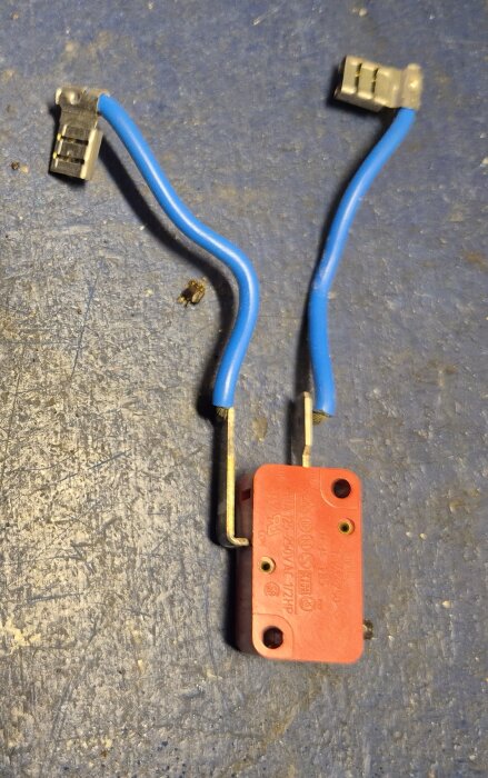 Röd mikrobrytare med två blå kablar liggande på en blå yta, 28x16mm och 10mm tjock.