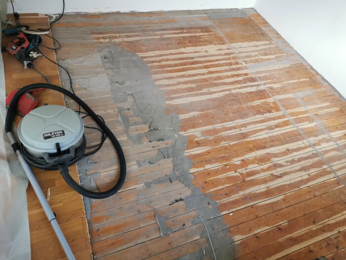 Gammalt plankgolv delvis täckt av flytspackel och lim med slipmaskin och verktyg bredvid.