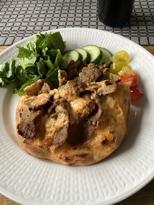 Tallrik med kebabpinza toppad med kött och ost, omgiven av sallad, gurka, tomat och paprika på ett bord.