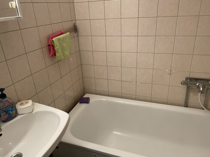 Badrum med vitt kakel, handfat, och badkar med armatur. Två handdukar hänger på vägg. Golvbrunn finns ej synlig.