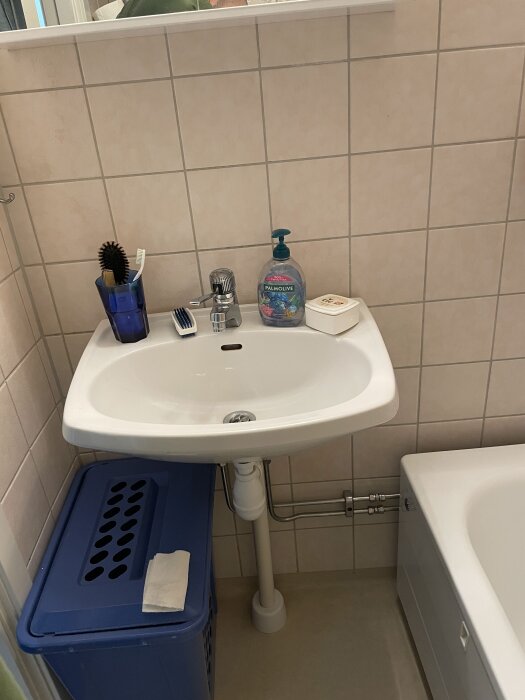 Handfat i badrum med tvål och tandborste på kanten, blå tvättkorg under och synliga rör. Delen av ett vitt badkar syns till höger.