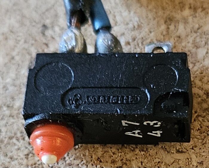 En svart elektrisk komponent med metallförbindelser och en röd knapp, text svår att tyda. Komponenten sitter intryckt i kassetten.