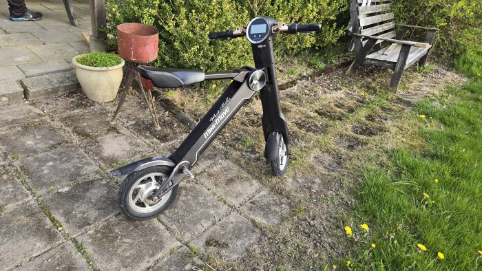 Svart elektrisk sparkcykel med sadel står på stenlagd uteplats bredvid kruka och träbänk, omgiven av grönska och maskrosor.