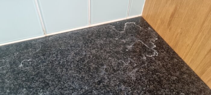 Vita fläckar på en mörk diskbänk nära kakelvägg och träpanel, troligen mineralavlagringar från vatten.