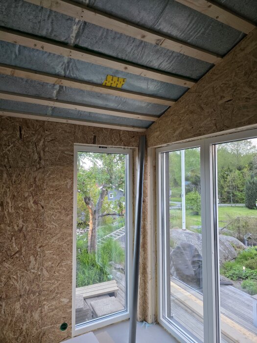 Ett rum under konstruktion med isolering i taket och spånskivor på väggarna, stora fönster och utomhusvy på trädgård med träd och gräs.