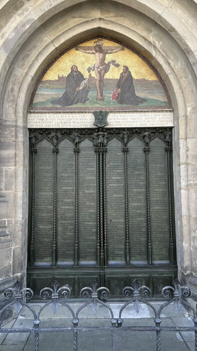 En utsmyckad kyrkport i Wittenberg med text i koppar och en mosaik av Jesus på korset med två figurer intill.