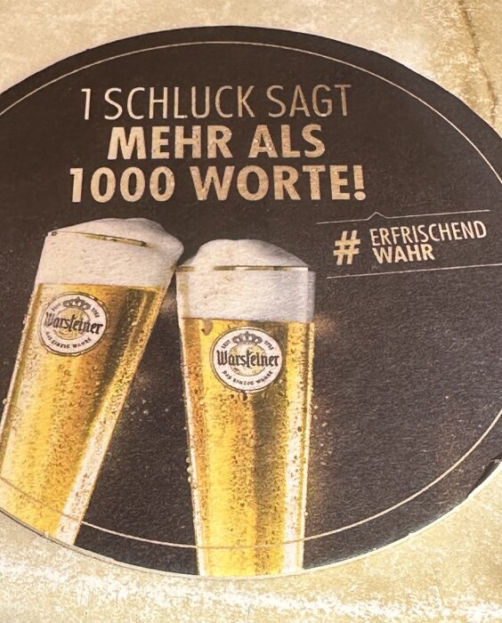 Två ölglas med Warsteiner-logga och text "1 Schluck sagt mehr als 1000 Worte! #Erfrischend Wahr" på en ölbordstablett.