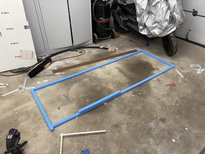 Blå ram på garagegolv med verktyg och material omkring, bredvid står en delvis övertäckt skoter.