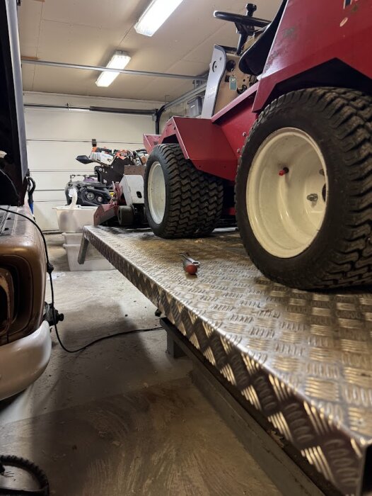 Röd åkgräsklippare på metallramp i garage med verktyg på rampen, omgiven av bilar och verktyg i bakgrunden.