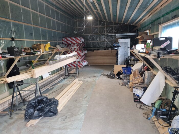 Trästomme och material i ett rum under byggnation, med verktyg och utrustning sorterade på hyllor längs väggarna.