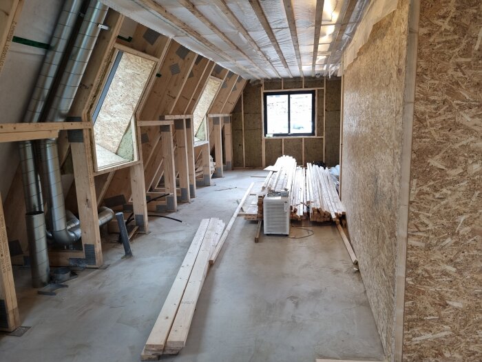 Ett renoverat rum med träramar, rörsystem och staplade träplankor. Vindsisolering och fönster installerade.
