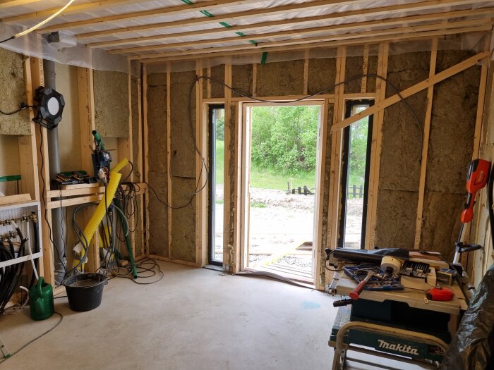 Renoveringsarbete i ett rum med synlig isolering, byggmaterial och verktyg spridda. Öppen dörr visar utsikt mot en grön trädgård.