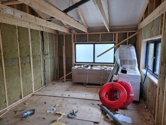 Rum under renovering med isolering i väggarna, staplade byggmaterial, verktyg och en röd slangrulle mot väggen.