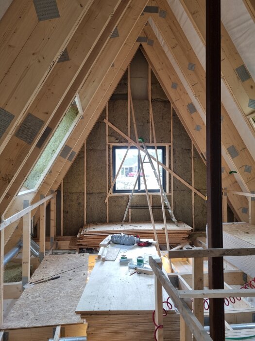 Byggarbetsplats i ett takutsprång med träbjälkar och isolering, verktyg och material ligger på träbord. Fönster i bakgrunden.
