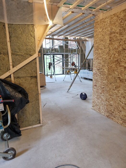 Inomhusbild av ett hus under byggnation med synlig isolering, träreglar och byggställning i bakgrunden.