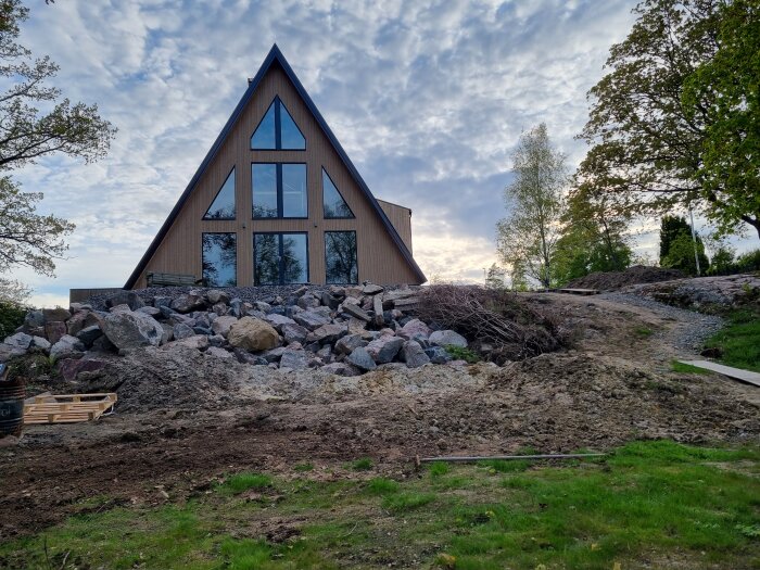 Triangulärt hus på bergig tomt med jord och gräsfläckar i förgrunden, omgiven av träd under molnig himmel.