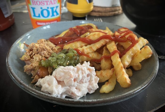 Tallrik med pommes, korv, räksallad, bostongurka, rostad lök, ketchup och senap. Bakgrund med burk rostad lök och ketchupflaska.