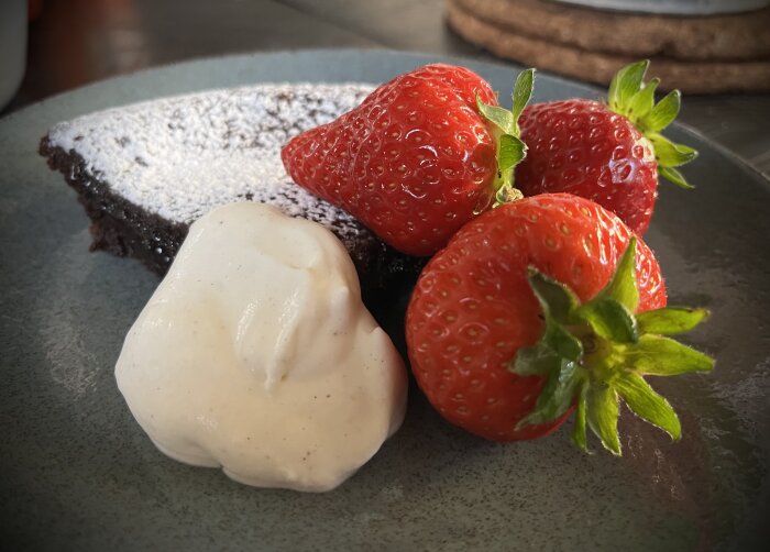 Pudrad kladdkaka med en klick vaniljgrädde och färska svenska jordgubbar på en tallrik.