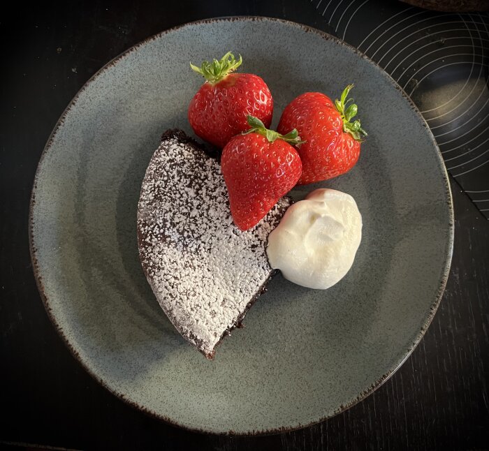 Pudrad kladdkaka serverad med tre jordgubbar och en klick vaniljgrädde på en tallrik.