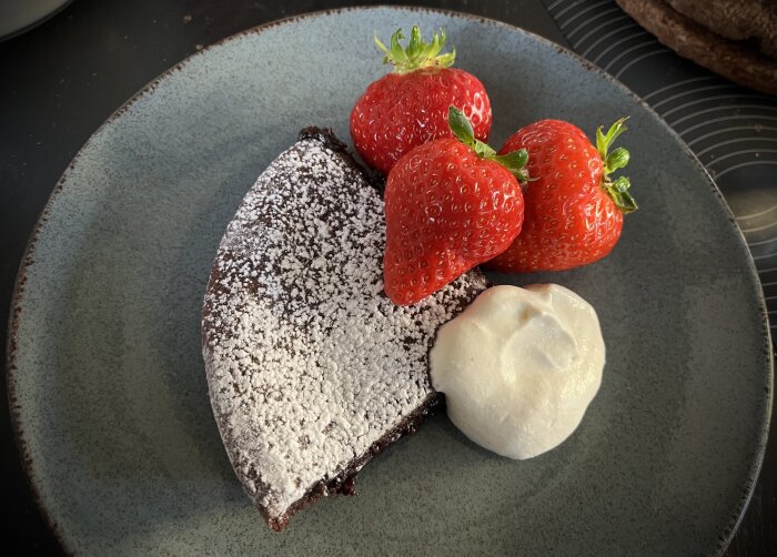 Pudrad kladdkaka på tallrik med tre svenska jordgubbar och en klick vaniljgrädde.
