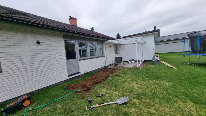 Grävda marken vid hus för ny trall och borttagen häck. Spade, handske och gräsklippare syns på gräsmattan.