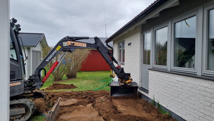 Grävmaskin tar bort matjord och gräs intill en byggnad för att förbereda för en ny altan, med en röd byggnad i bakgrunden.