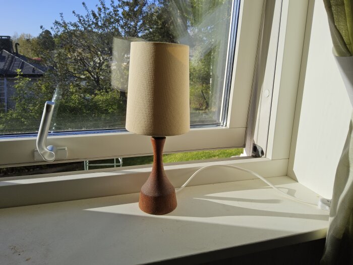 Fönster med utvändigt liggande fönsterbåge, öppnat uppåt. Skrivbordslampa med träfot står på fönsterkarmen, utsikt mot trädgård och hus.