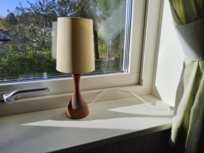 Fönster med silverfärgat handtag, en bordslampa med träfot och beige skärm på fönsterbrädan, gröna gardiner i bakgrunden.