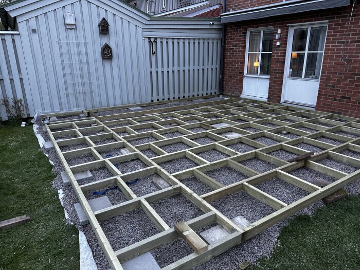 Träram för altan på grusgrund framför rött tegelhus och vitt plank, påbörjad byggnation av en 6x6 meter trall.
